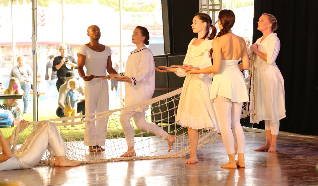 Image of 6 dancers dressed in white, four holding wooden tennis rackets and the others holding a tennis court net. 