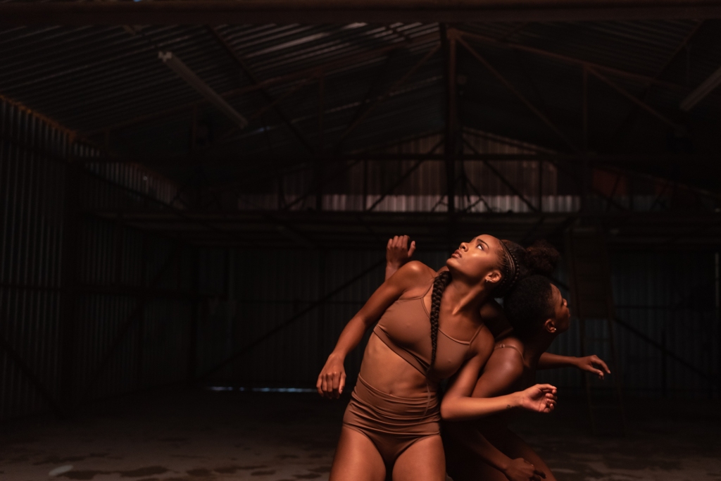 Three dancers connected and leaning on each other in a warehouse.