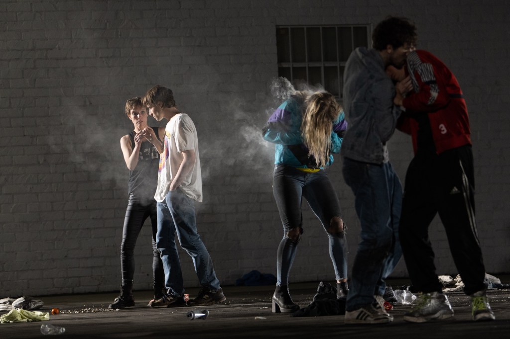Dancers standing and interacting through touch while a solo dancers hunches with smoke coming out of her jacket. 