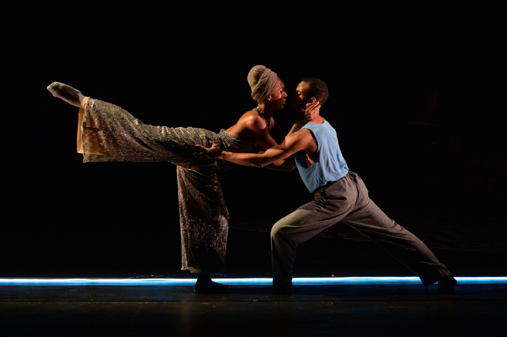 Woman and man dancing together. Woman is in an arabesque with a silver jumpsuit and headwrap. Man is lunging with a blue tank top and grey trousers. 