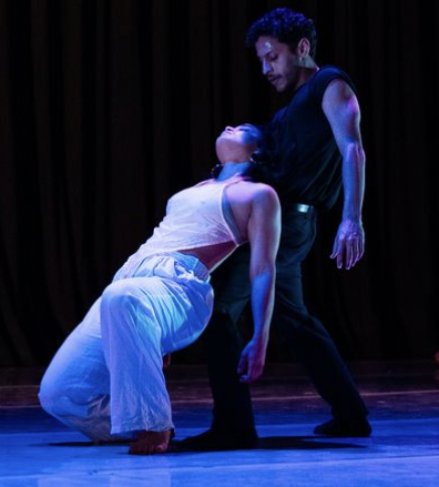 Female dancer dressed in light colors leaning backward against the torso of the standing male dancer dressed in black. 