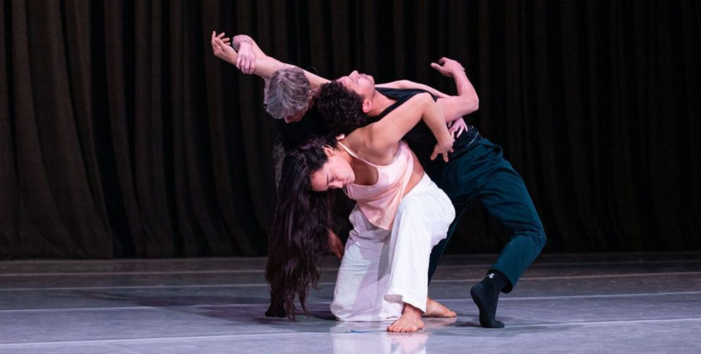 Three dancers huddled together into a unified shape with the male dancer arching backward over them. 