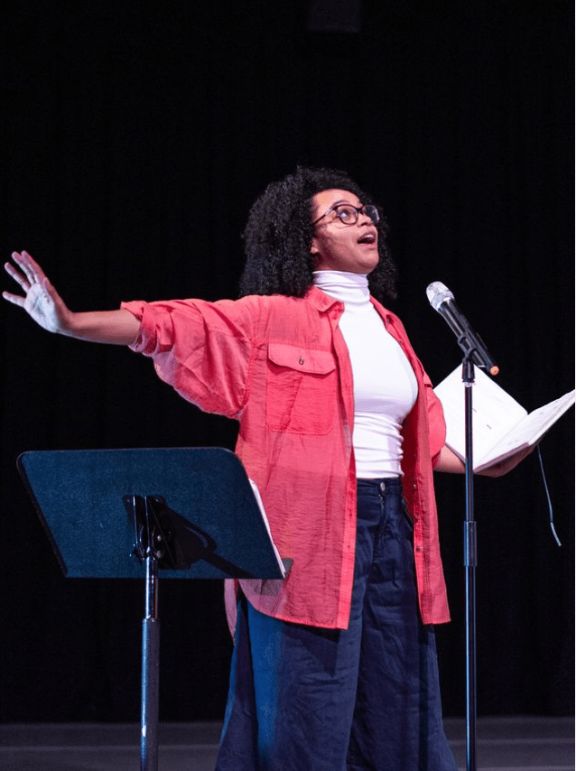 Female dancer wearing a pink button down shirt speaking into microphone on a stand with her arms reaching outward and looking forward and up.
