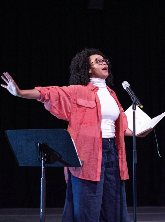 Female dancer wearing a pink button down shirt speaking into microphone on a stand with her arms reaching outward and looking forward and up.