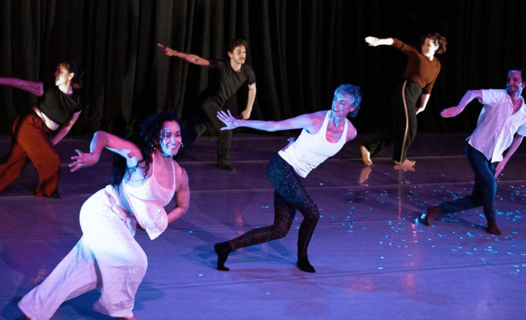 Six dancers in casual rehearsal clothes reaching to the right side while smiling. 