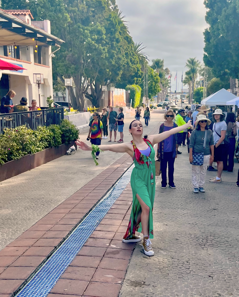 Dancer in teal and red dress reaching outward while dancing along a pedestrian zone. 