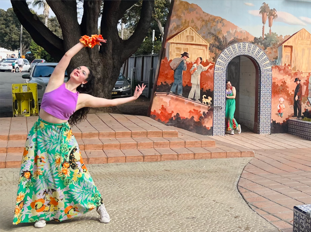 Dancers performing outside in long colorful skirts. One stands in the foreground with arms overhead and one is poised in a nearby archway. 