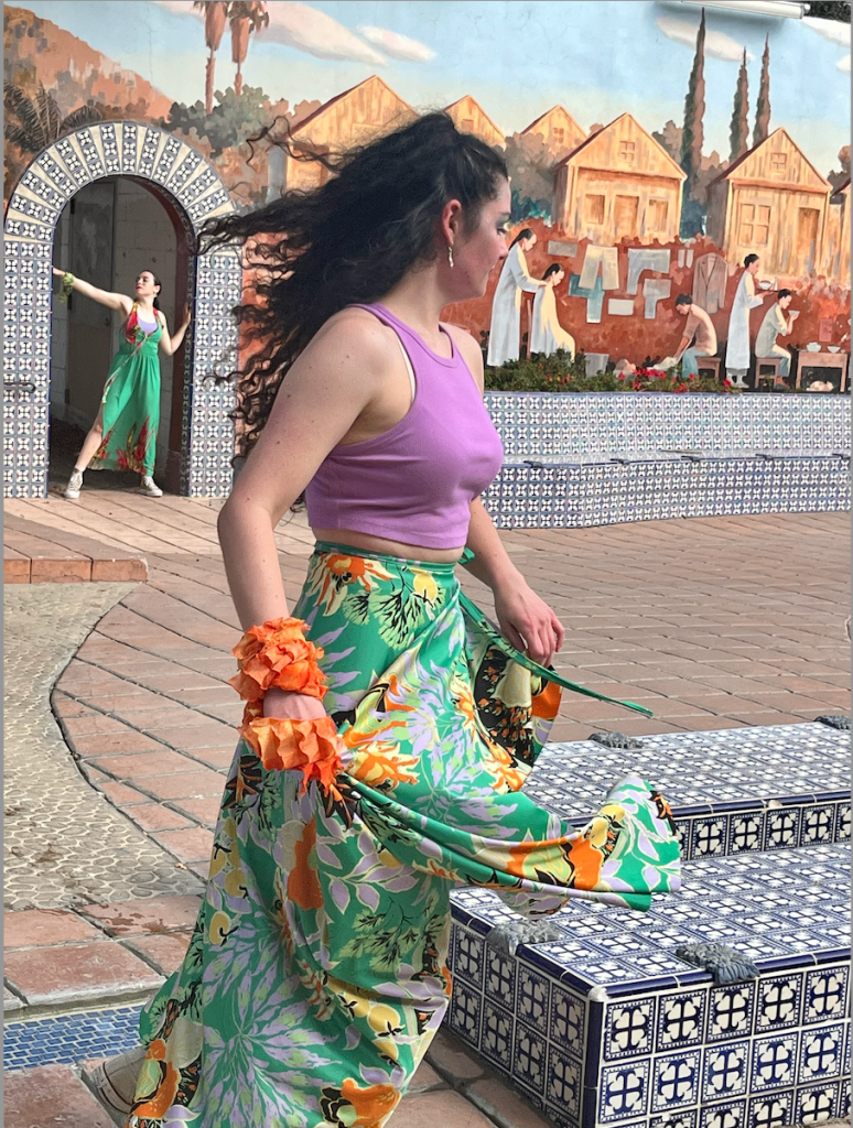 Dancers in long skirts dancing by a fountain and against a tiled wall. 