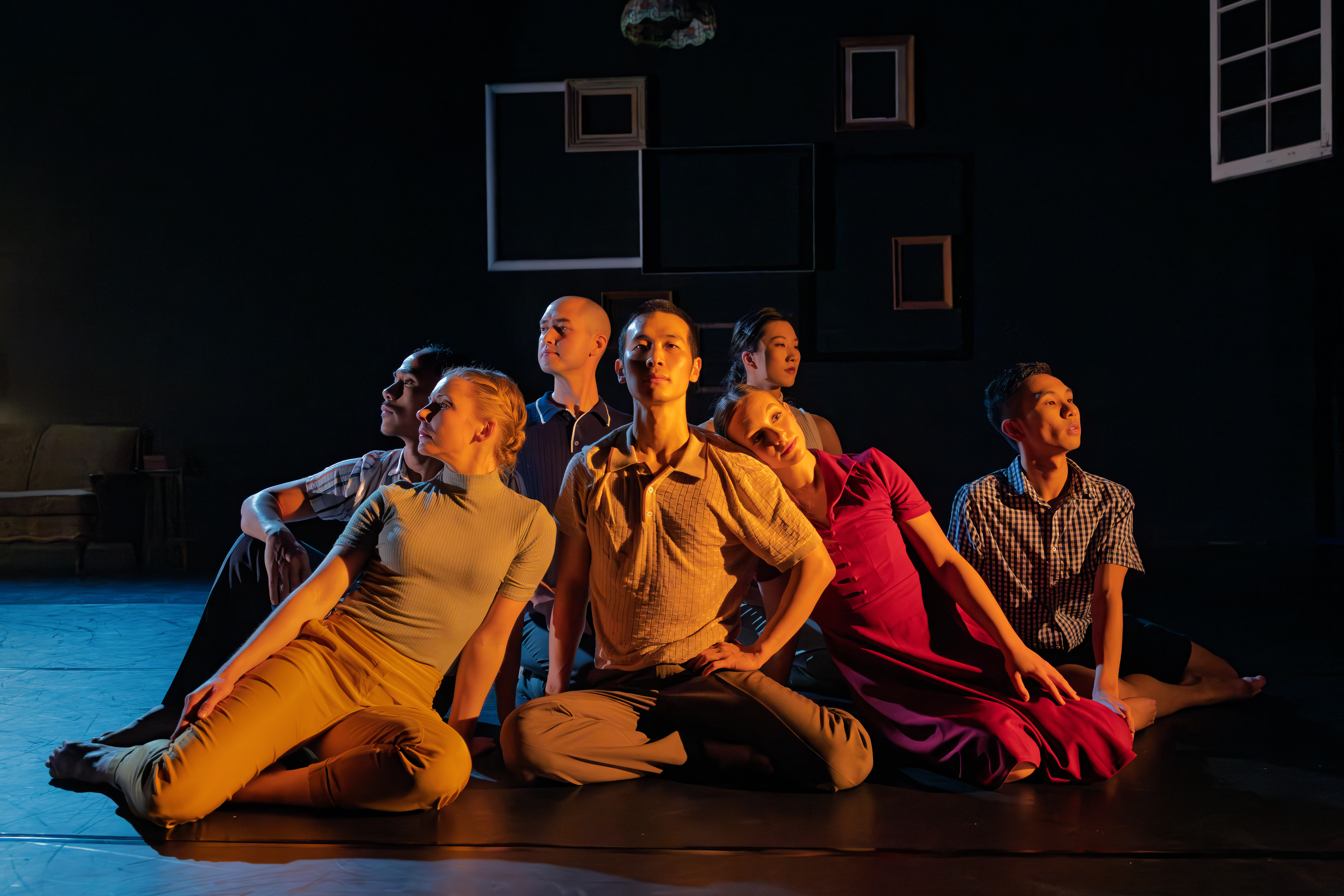 Cast members of home seated in uplifted postures on the ground in a clump looking in various directions in warm amber light.